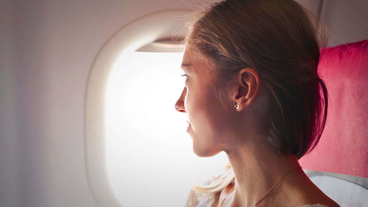 Woman with blonde hair in a ponytail sitting in an airplane seat looking out the window in soft golden light | running away abroad | fear of settling down expat | international therapy for expats | England | Netherlands | France