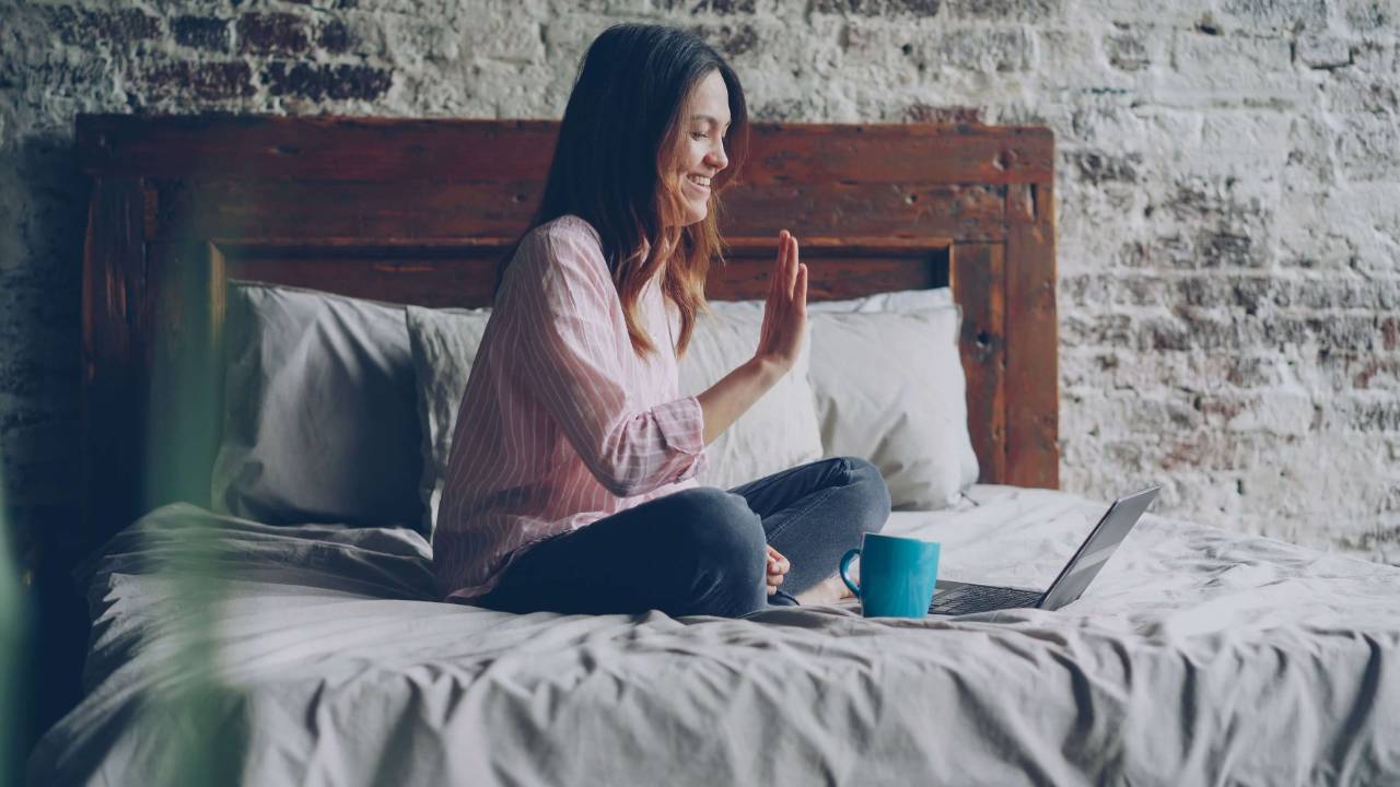 Woman in a pink striped shirt sitting cross legged on a bed smiling and waving at a laptop with a blue mug beside her | therapeutic coaching online | international trauma therapist | private pay therapy alternative | New York City | Chicago | Los Angeles