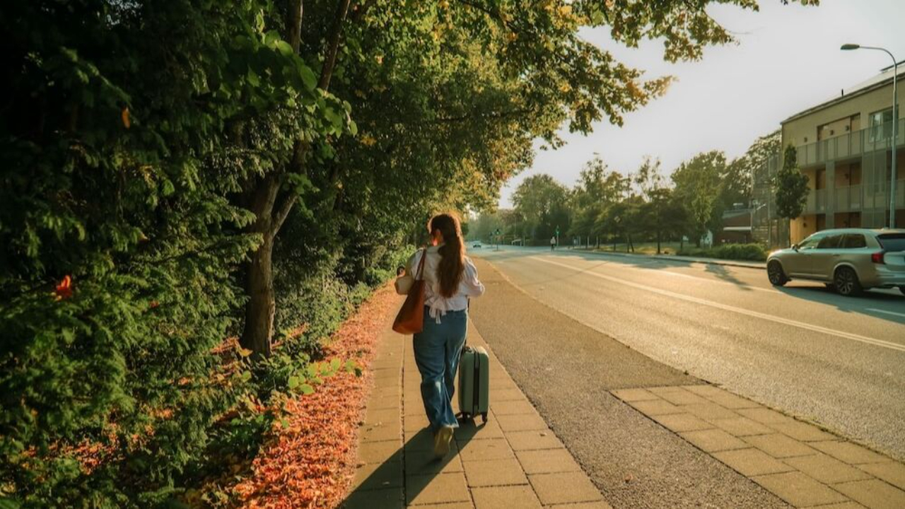 Woman with long red hair walking along a sunny autumn pavement pulling a suitcase beside a tree lined road | international therapy and coaching | expat attachment | globally mobile relationships | Germany | Netherlands | Scotland