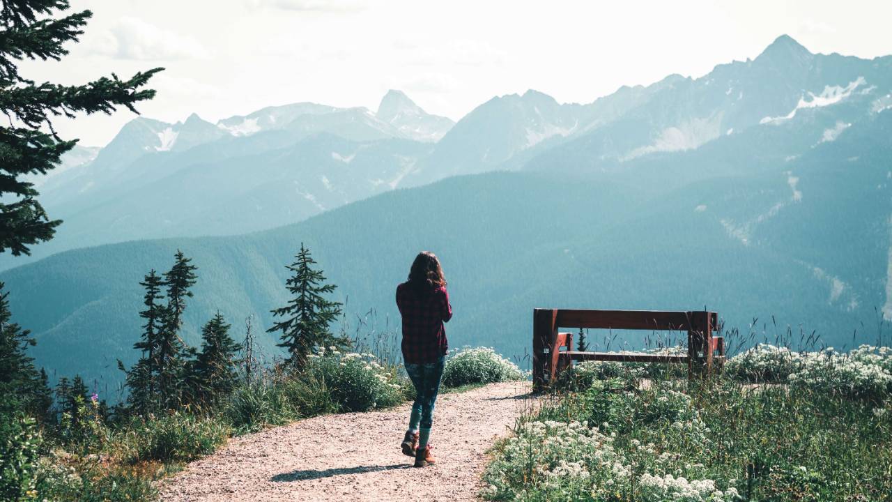 Woman in a burgundy jacket walking alone on a gravel mountain path toward a wooden fence overlooking misty snow capped peaks | somatic energy therapy | therapeutic coaching online | international trauma therapist | Seattle | Los Angeles | New York City
