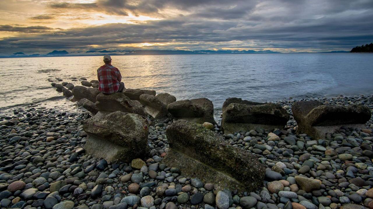 Man in a red plaid shirt and cap sitting alone on a rocky shoreline looking out over a calm ocean at a dramatic cloudy sunset | parts work training for therapists | international trauma therapist | therapeutic coaching | New York City | Chicago | Los Angeles