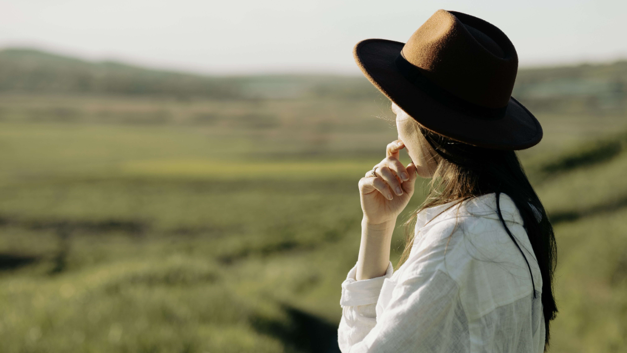 Woman in a wide brim black hat and white linen shirt looking out pensively over a green open landscape | expat social connection | transient life loneliness | international therapist | Scotland | Canada | France