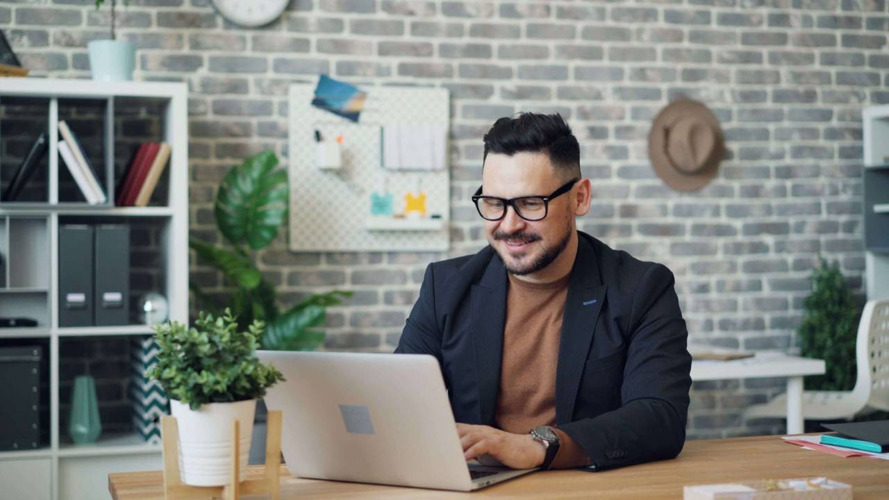 Man with glasses and a dark blazer smiling while typing on a laptop at a wooden desk in a creative brick walled office with plants | executive coaching | therapeutic coaching | international trauma therapist | Chicago | Los Angeles | Seattle