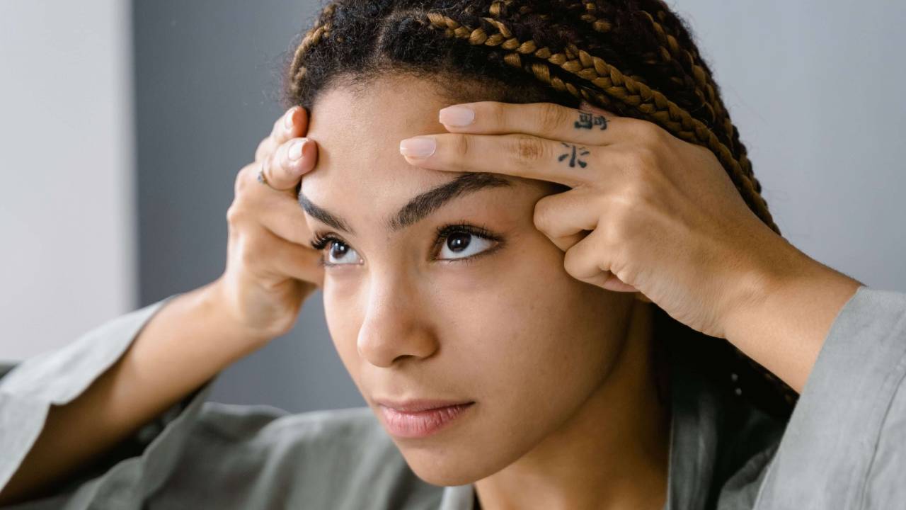 Woman with braided hair and tattoos pressing her fingers to her forehead and temples with a focused expression against a grey background | tapping for trauma | tapping therapist | international trauma therapist | New York City | Chicago | Los Angeles