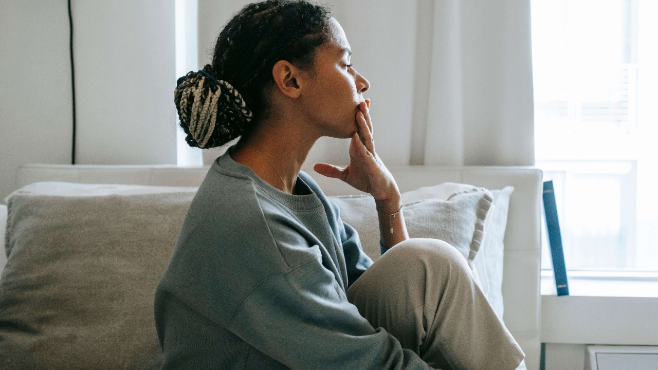 Woman with braided hair in a grey sweatshirt sitting on the floor leaning against a sofa with her hand to her mouth in quiet reflection | high functioning trauma | burnout trauma professionals | international trauma therapist | South Africa | Canada | Netherlands