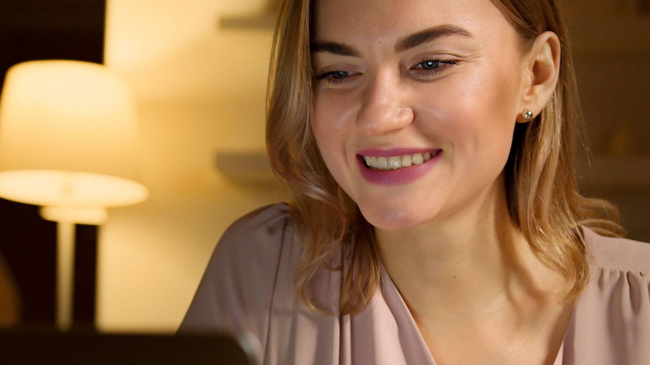 Woman in a pink wrap top smiling warmly at a laptop screen in a softly lit room with a lamp in the background | EMDR therapy online | virtual EMDR | international trauma therapist | England | Netherlands | Canada