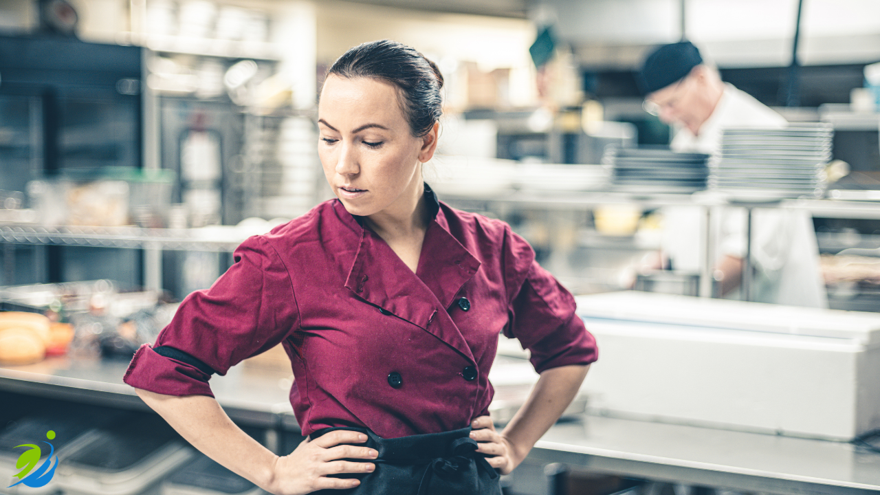 Restaurant manager standing in a busy kitchen with hands on hips, observing operations and identifying potential safety issues in a fast-paced environment.