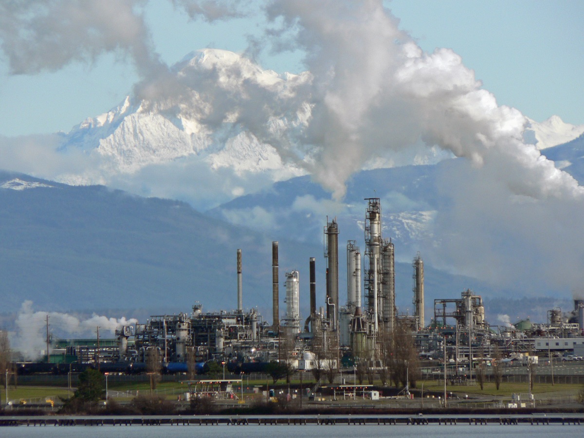 An oil refinery seen across water in daylight: tall distillation columns, stacks, pipe racks and process units, with a row of rail tank cars at the base.