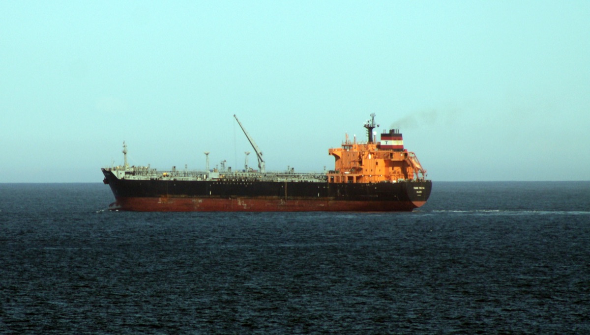 A tanker under way in open sea under a hazy sky, side-on: black hull, red lower hull, orange superstructure, deck pipework and one hose crane.
