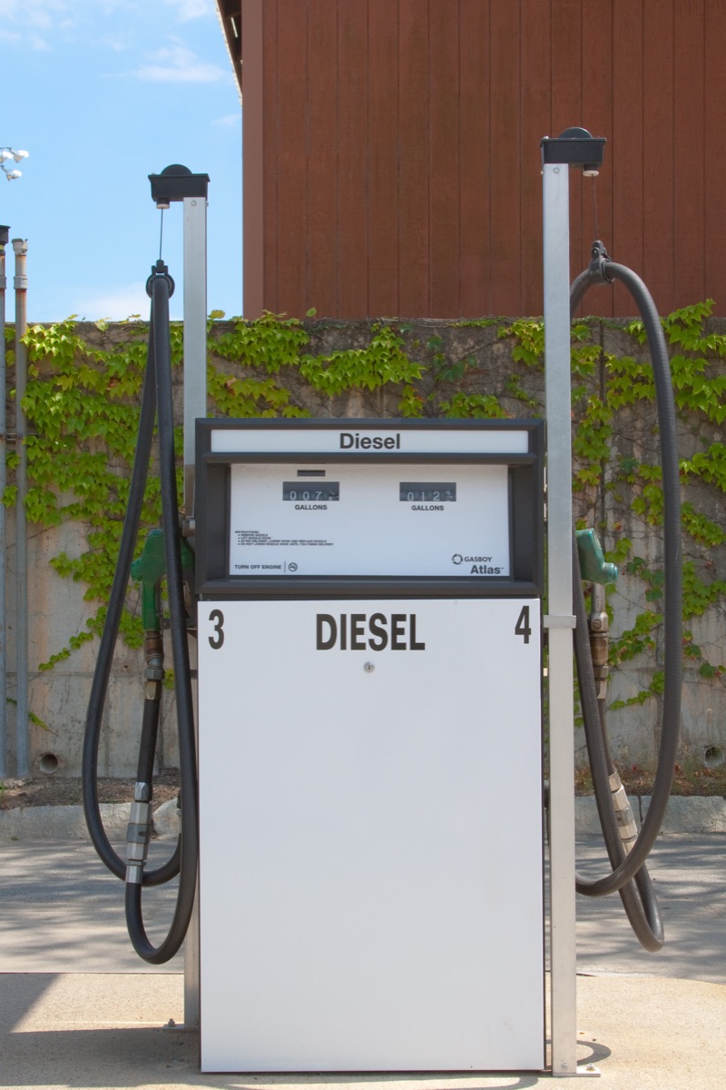 A plain white diesel dispenser in sunlight with two hoses and green nozzles.