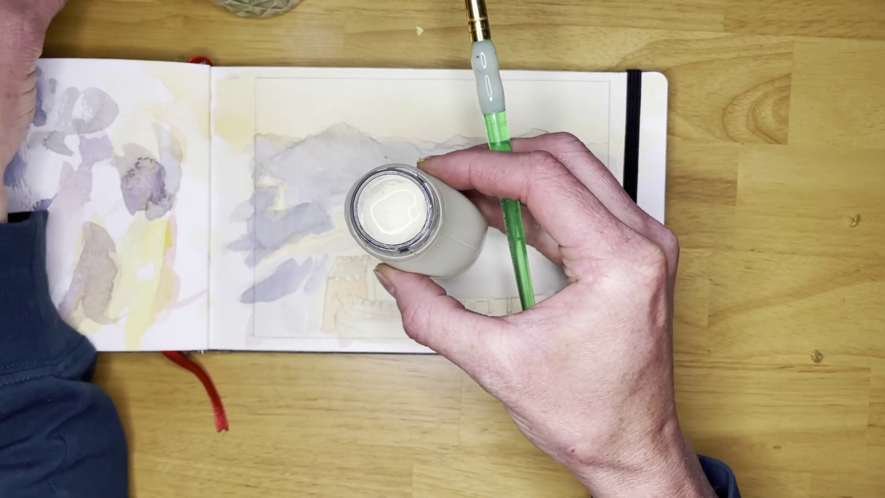 Artist Juliene holding a bottle of masking fluid above a watercolor sketchbook with a paintbrush nearby, demonstrating how to use masking fluid in watercolor to preserve clean white highlights.