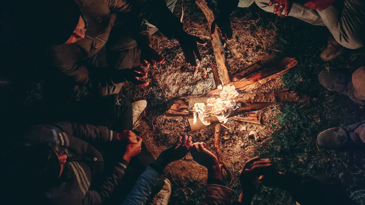 People gathered close around a small campfire at night, hands reaching toward the flames