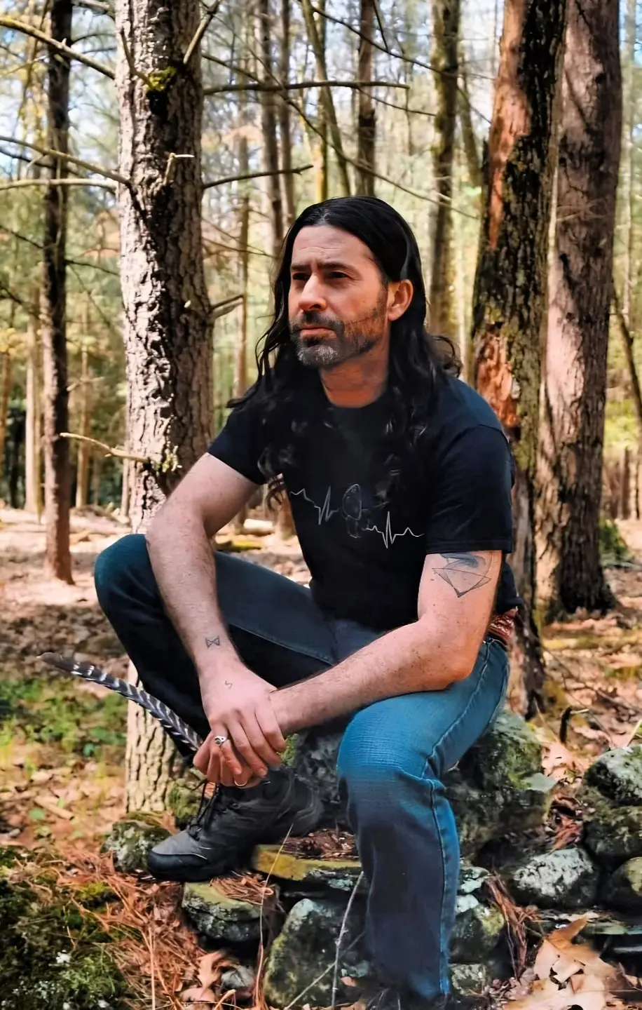Angell Deer, founder of Sacred Paths, sitting on a mossy stone wall in a pine forest, holding a feather