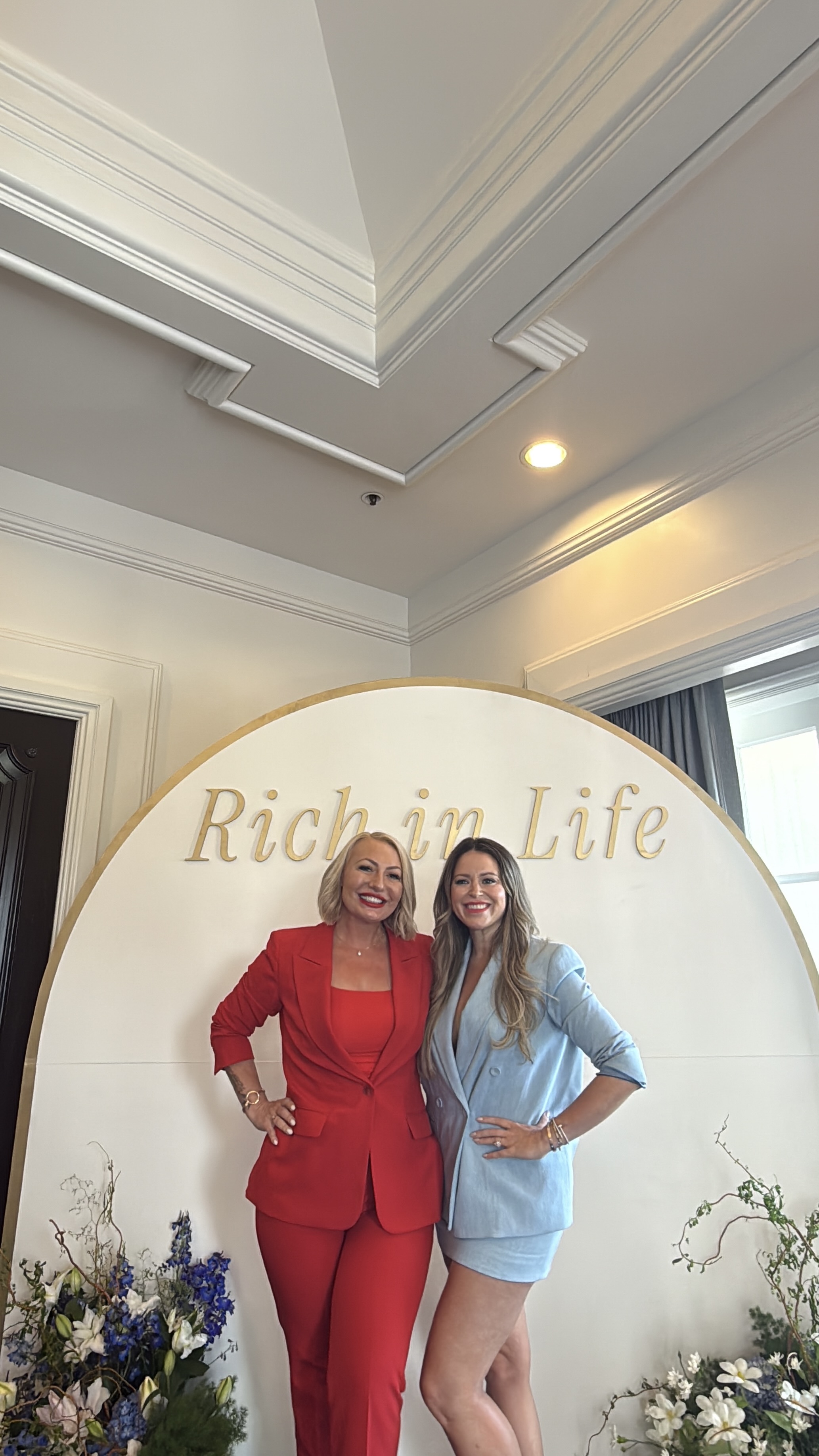 Miranda in a red suit with a chiropractic leader in front of a Rich in Life arch