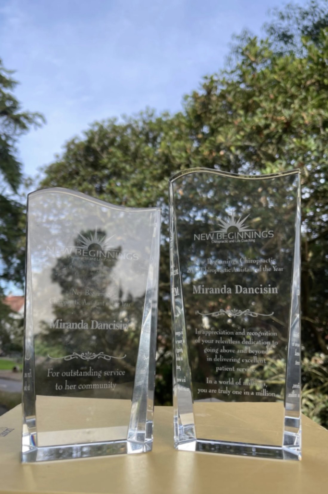 Miranda's two crystal New Beginnings Chiropractic Assistant of the Year awards, engraved with her name, outdoors in front of trees