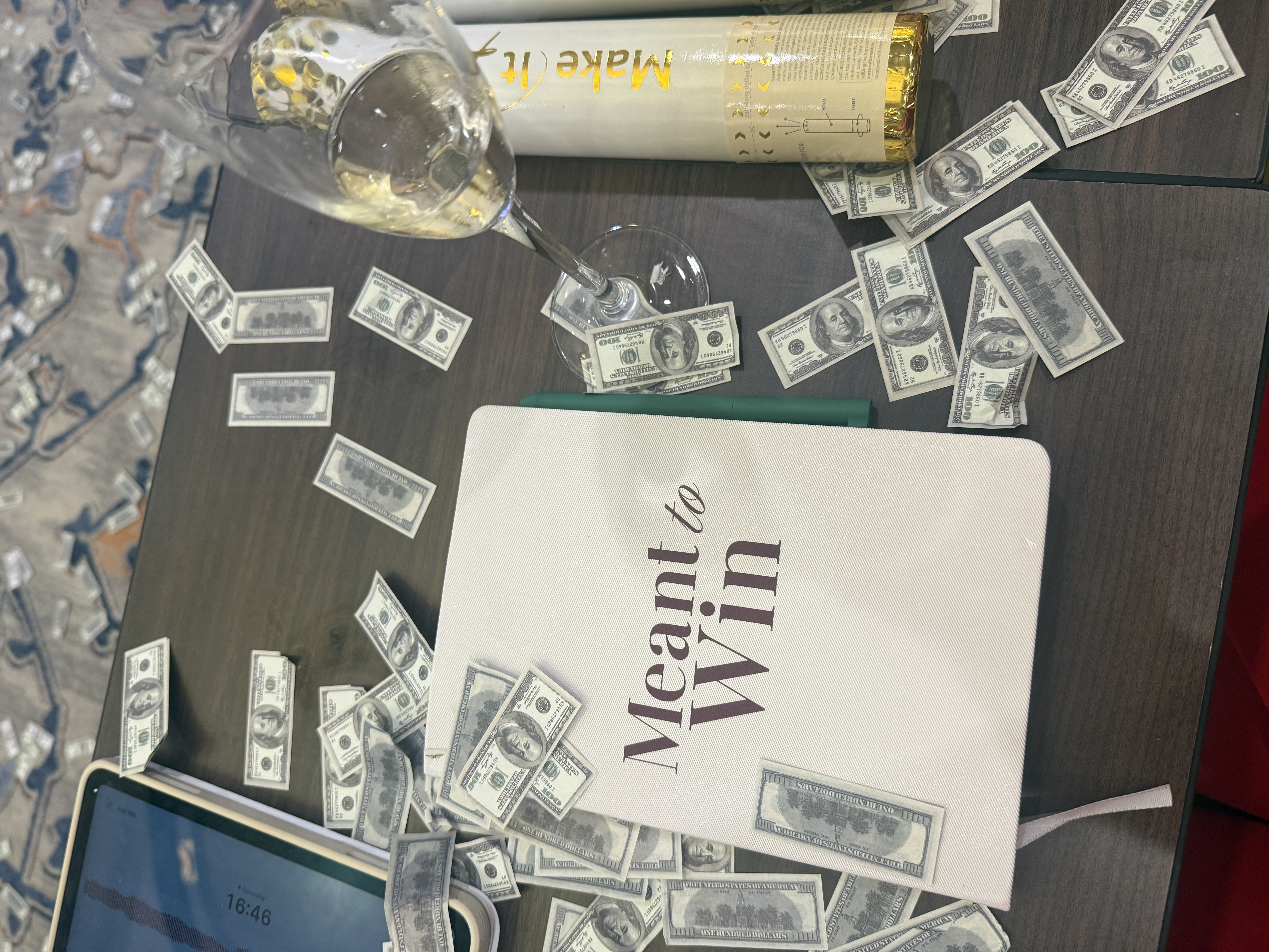 A Meant to Win journal on a table covered in play money next to a bottle of champagne