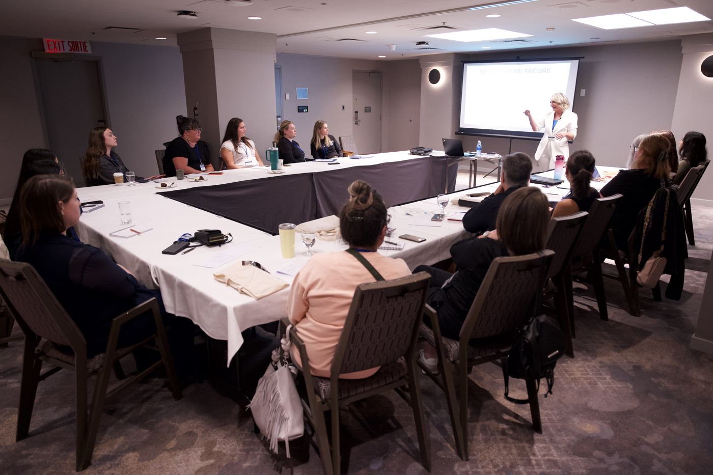 Miranda teaching a room of C.A.s at a U shaped table, with Vital Word number 5, SECURE, on the screen