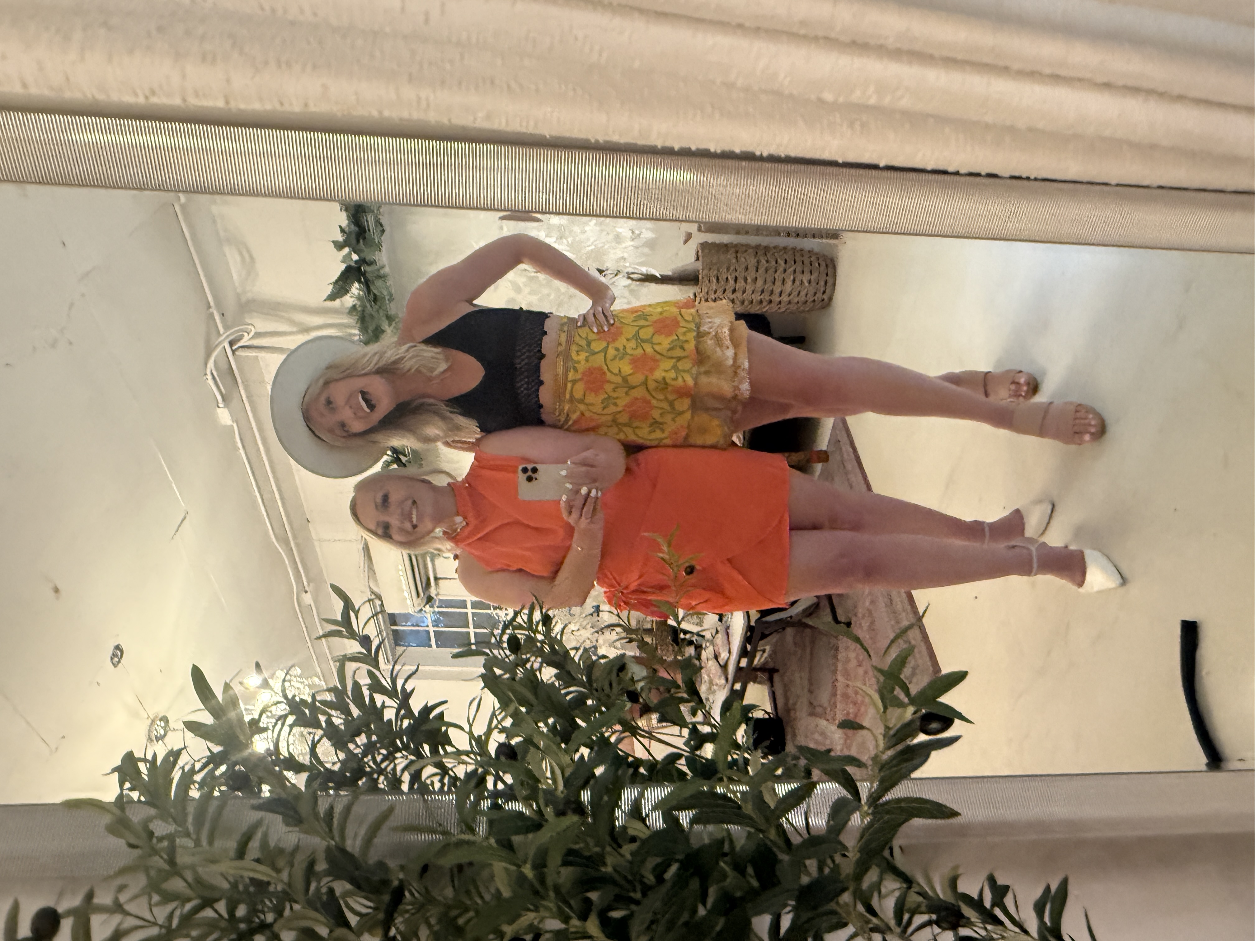 Miranda in orange taking a mirror selfie with a friend in a wide brim hat