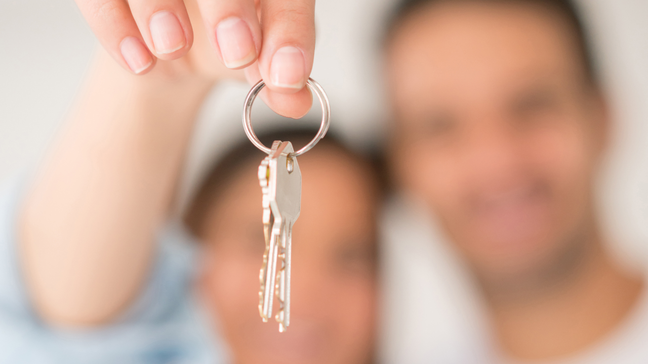 A close-up of a hand holding up a set of house keys, with a blurry couple smiling in the background.