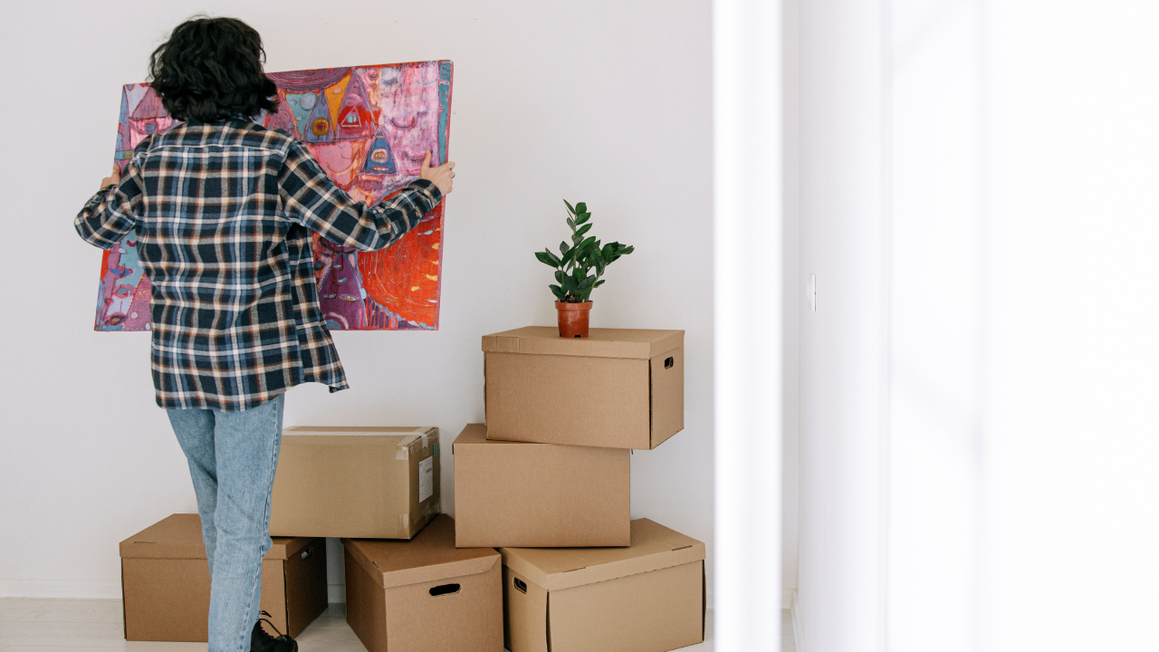 Person unpacking a vibrant artwork in a room filled with cardboard relocation boxes, illustrating the emotional transition of moving.