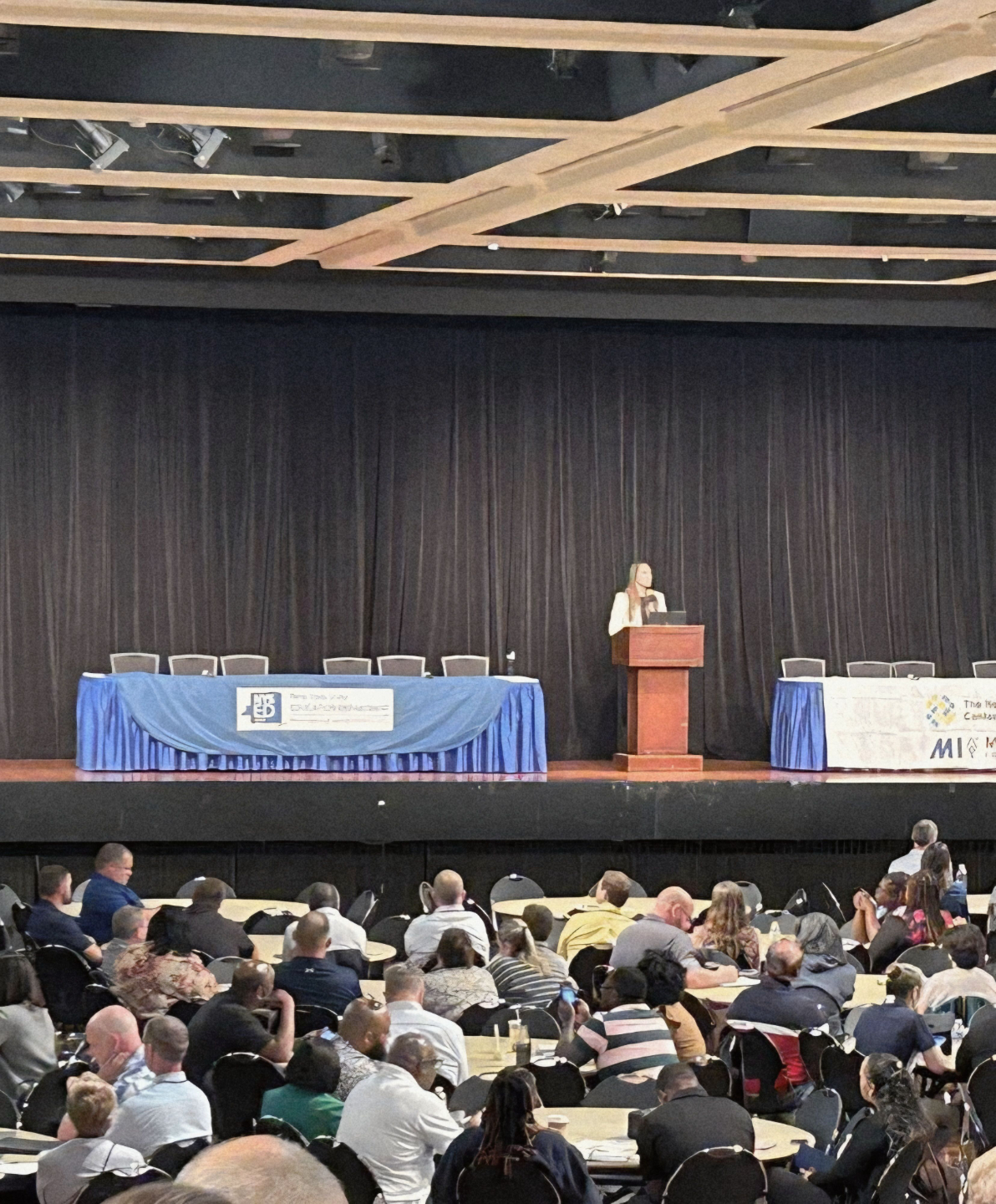 Christina Pate wearing a white blazer standing on stage behind a podium giving a speech with two long tables on both sides of the podium and people sitting in chairs at tables on the floor beneath the stage.