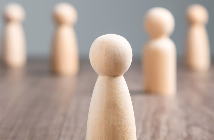 Wooden block shaped like a person with a round top for the head and bottle shape for the body standing in front of four same shaped blocks with a grey background and wooden floor.