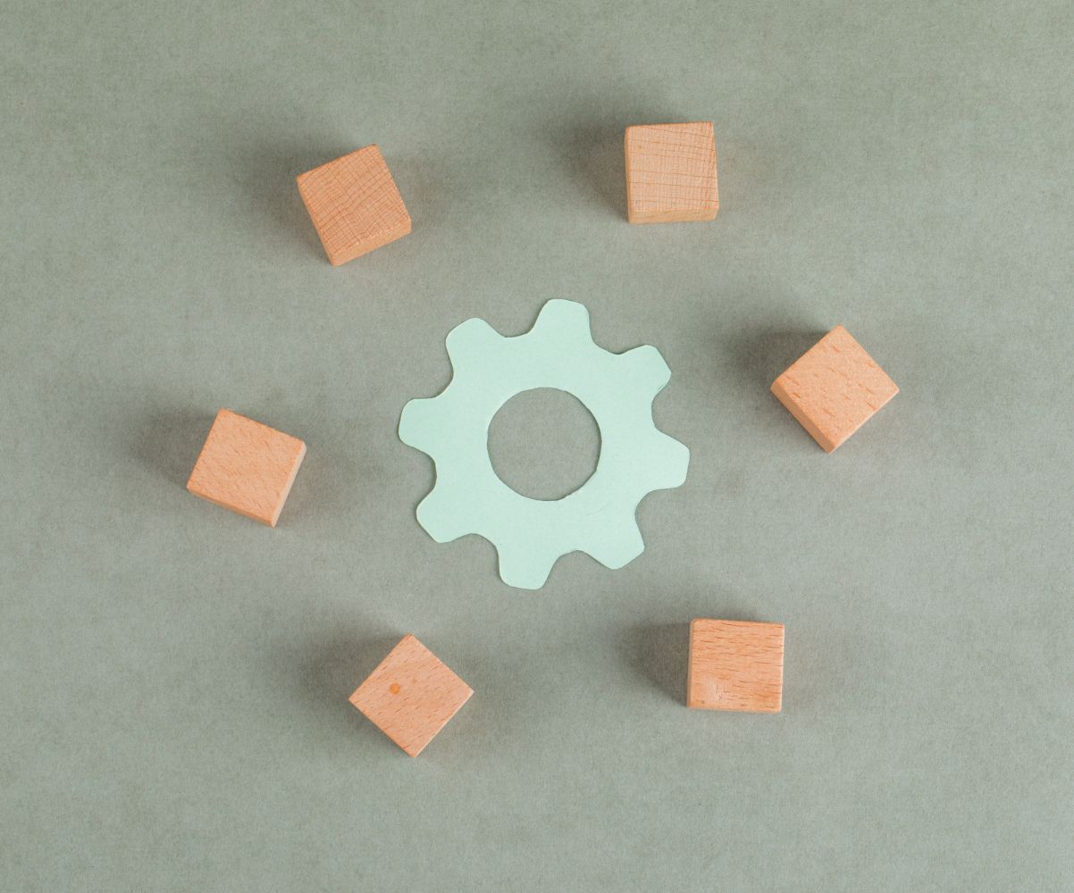Light green wooden gear-shaped object surrounded by six orange wooden blocks placed on a grey table.