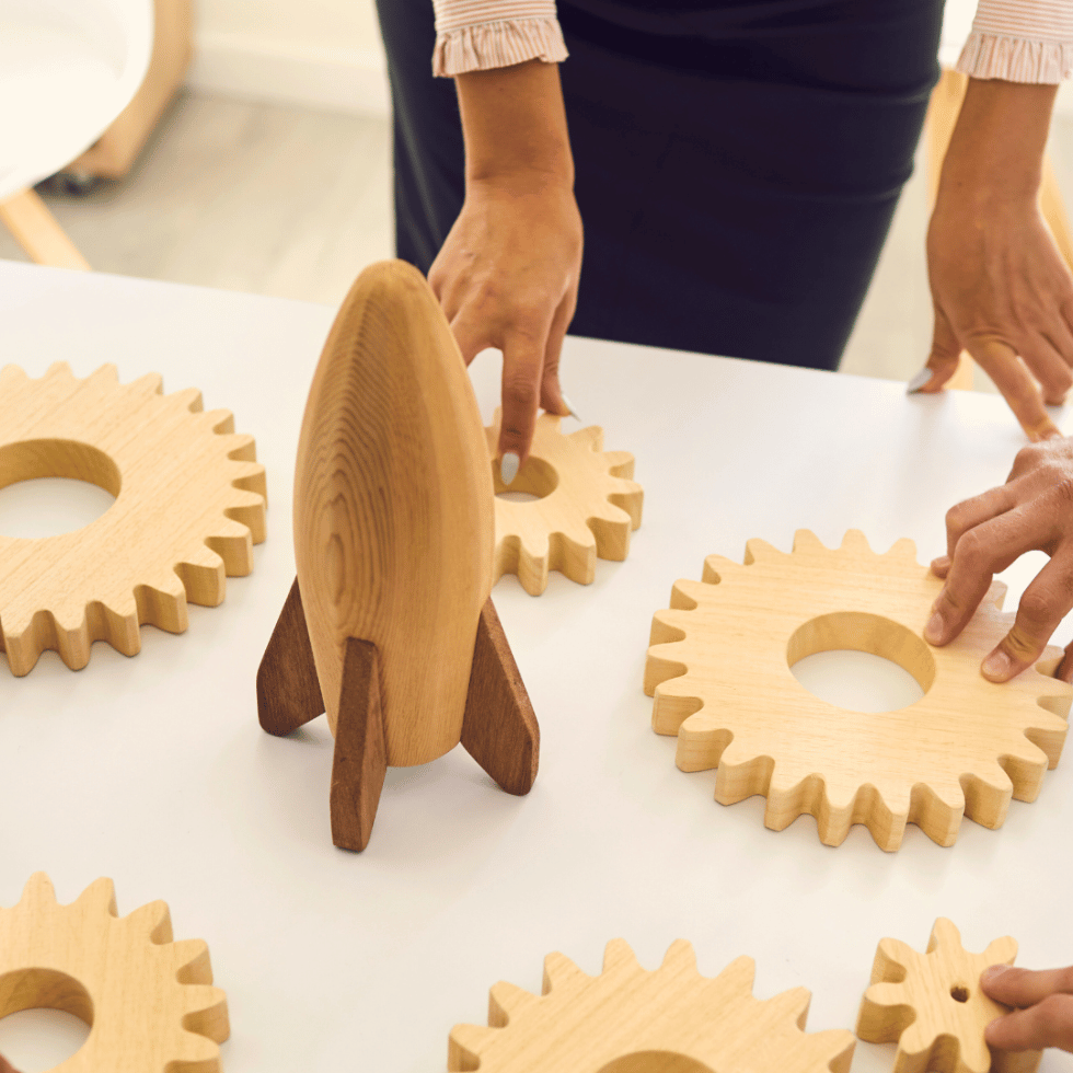 Two people’s hands touching wooden gear-shaped blocks on a table with a wooden rocket ship in the center of the table.