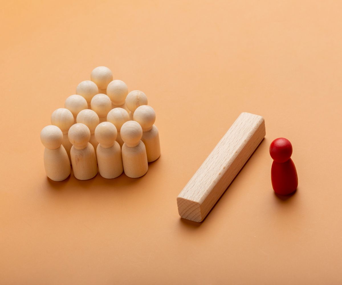 One red block shaped like a person standing behind a rectangular light-colored wooden block and a set of light-colored blocks shaped like people forming a pyramid shape facing the red block