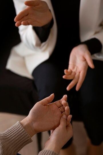 Two people sitting close face-to-face showing their hands only with palms up with one person wearing a white blazer and black pants and the other person wearing a brown sweater.
