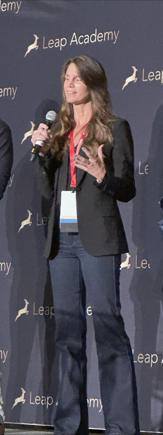 Christina Pate wearing black pants and black blazer standing on stage holding a microphone speaking with a background that says Leap Academy.