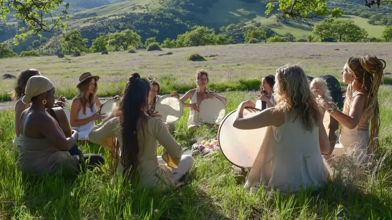 Krista Holland and women sitting in a circle in a field of grass playing frame drums width="1280" height="720"px