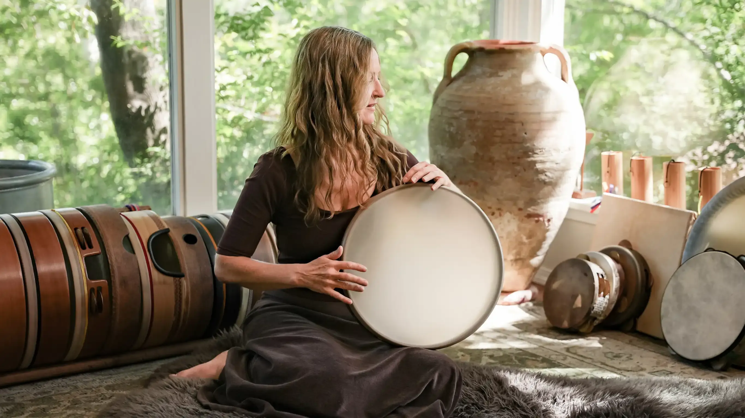 The Frame Drumming Academy founder Krista Holland wirh playing her frame drum in a room with many frame drums and instuments