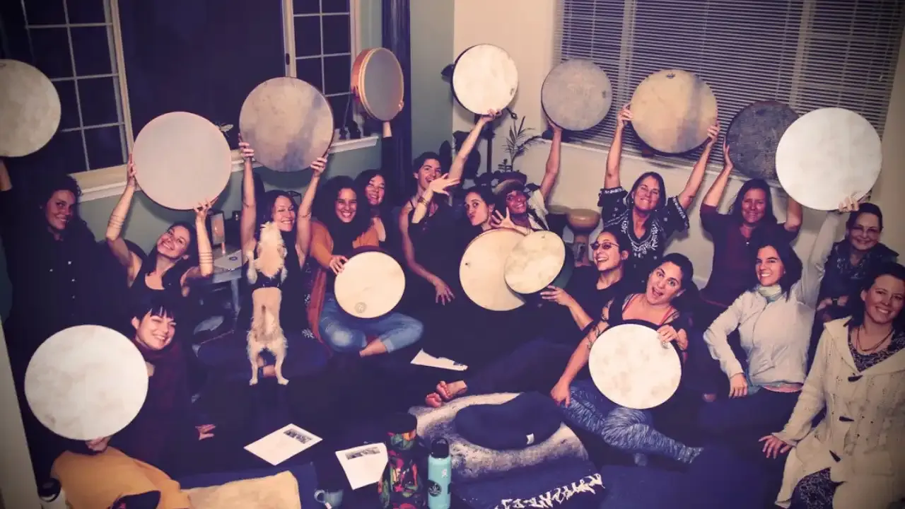 Krista Holland and Liron Meyuhas with a group of women sitting on the floor holding frame drum up in the air.