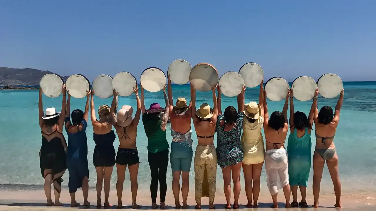 Krista Holland's frame drumming acadmey on retreat at elefonisi beach on Crete, Greece in a line of 12 women holding frame drums over their heads looking at the sea