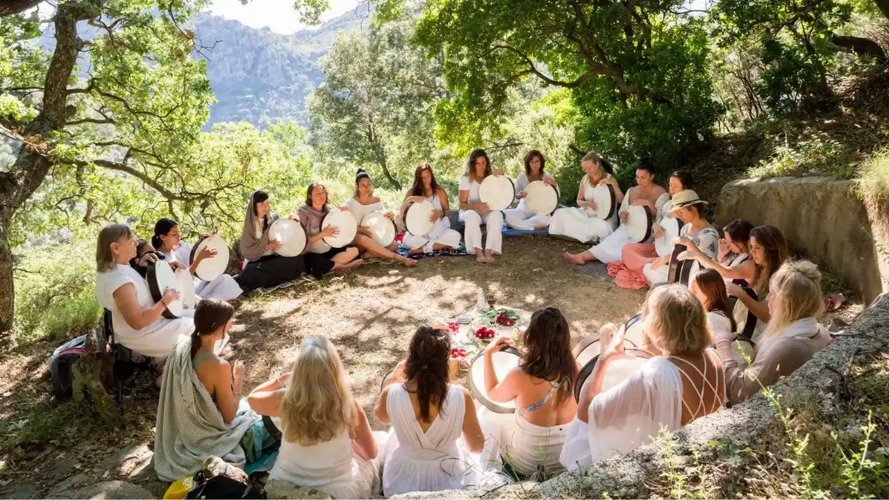 Krista Holland a a group of women wearing white dresses sitting in a circle playing frame drums under the shade of trees on the island of Crete, Greece.