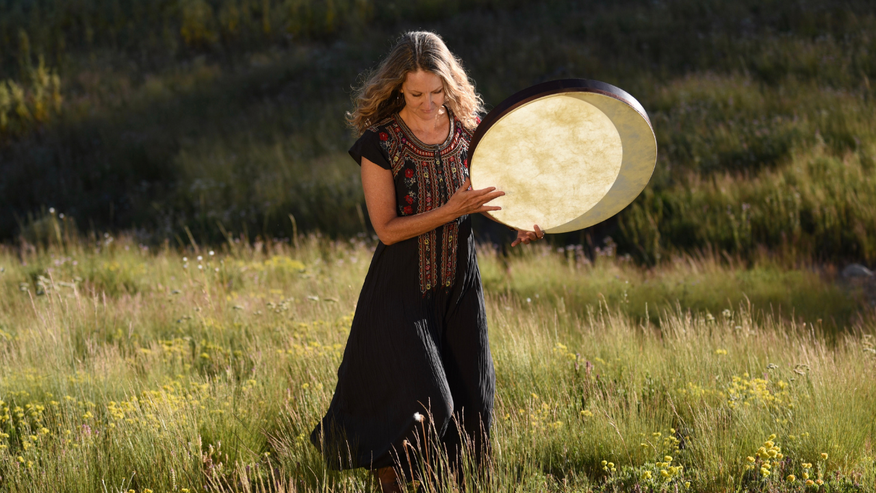 Krista Holland playing a frame drum in a meadow wearing an embroidered black Johnnie Was dress.