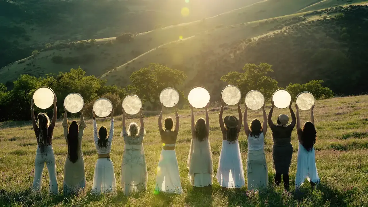 Krista Holland's Frame Drumming Academy with 10 women standing with their backs to the camera wearing white dresses holding frame drums over their heads illuminated by the sun width="1280" height="720"px