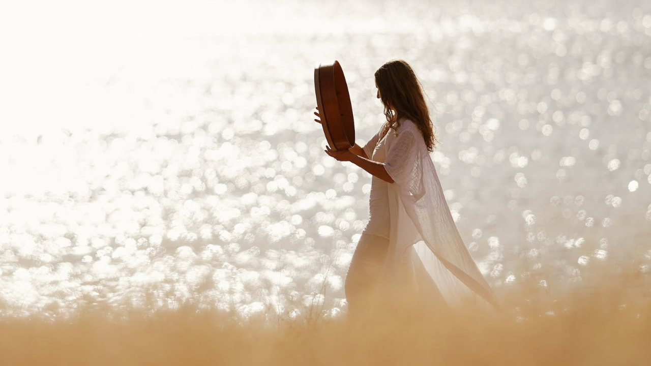 Krista Holland wearing a white dress playing a frame drum.