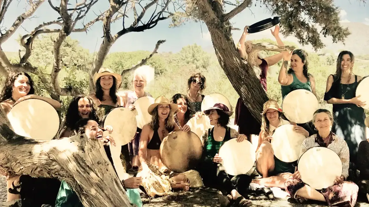 Krista Holland and a group of women sitting under carob trees at Elafonisi beach in Crete, Greece, playing frame drums 