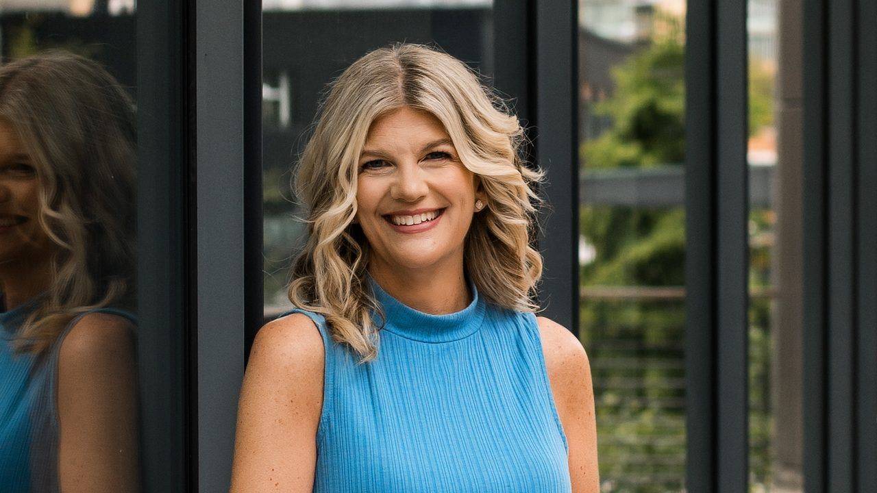 Stacey Salyer smiling in a blue sleeveless top beside a dark glass wall