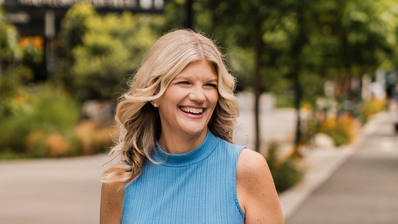 Stacey Salyer laughing in a blue sleeveless top on a tree-lined city sidewalk