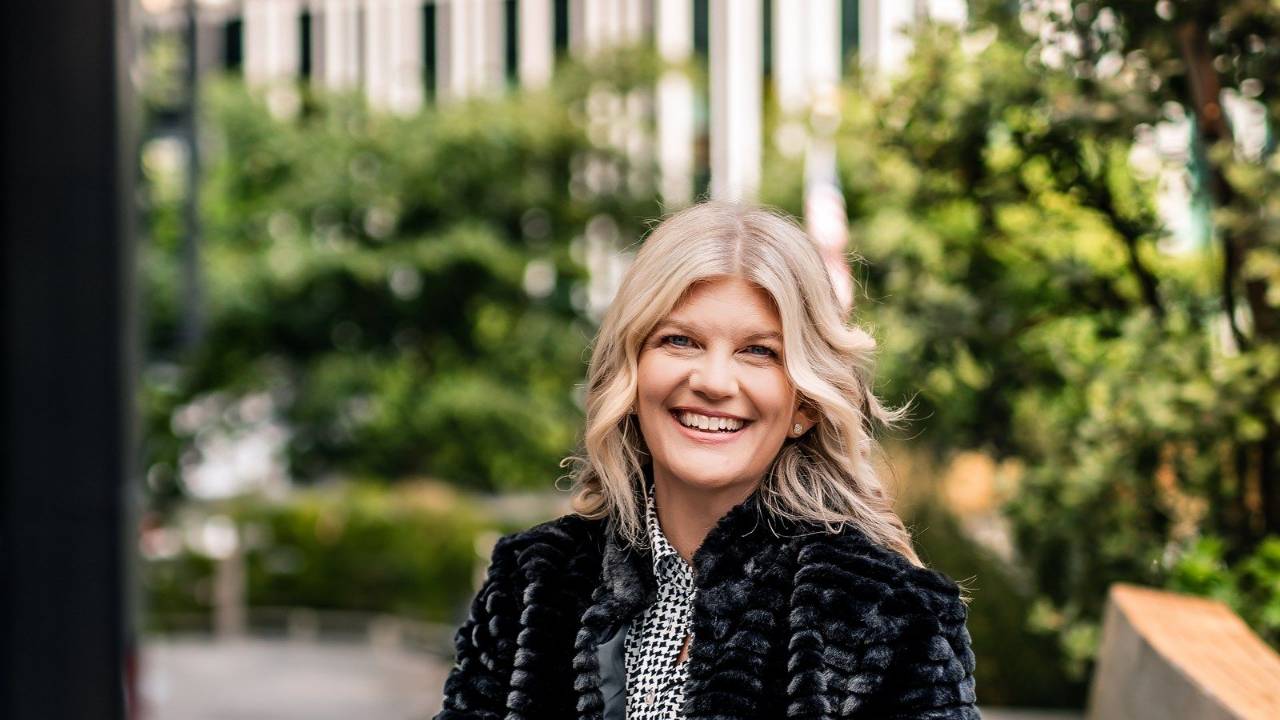 Stacey Salyer smiling warmly in a green outdoor courtyard, looking off camera