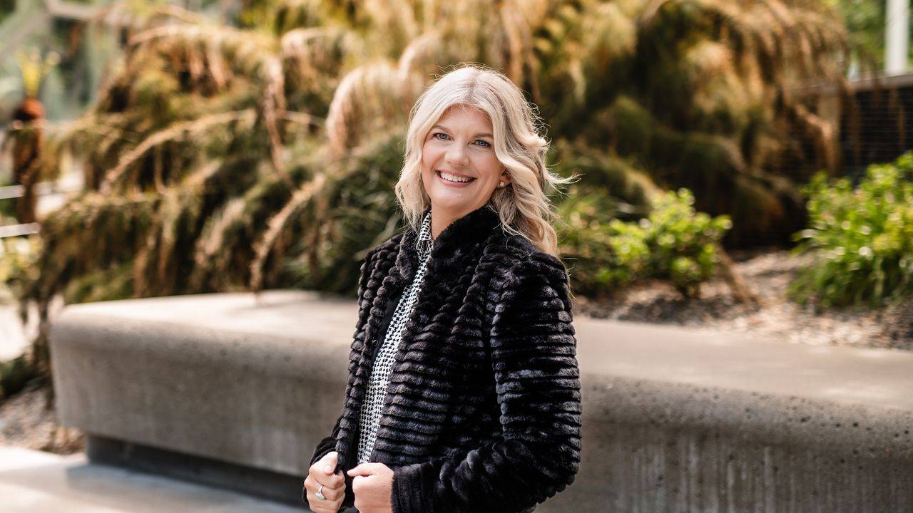 Stacey Salyer smiling in a black faux fur jacket outdoors beside a stone bench and grasses