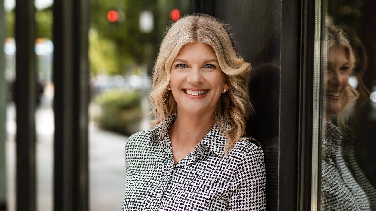 Stacey Salyer smiling in a black and white houndstooth blouse beside a dark glass wall