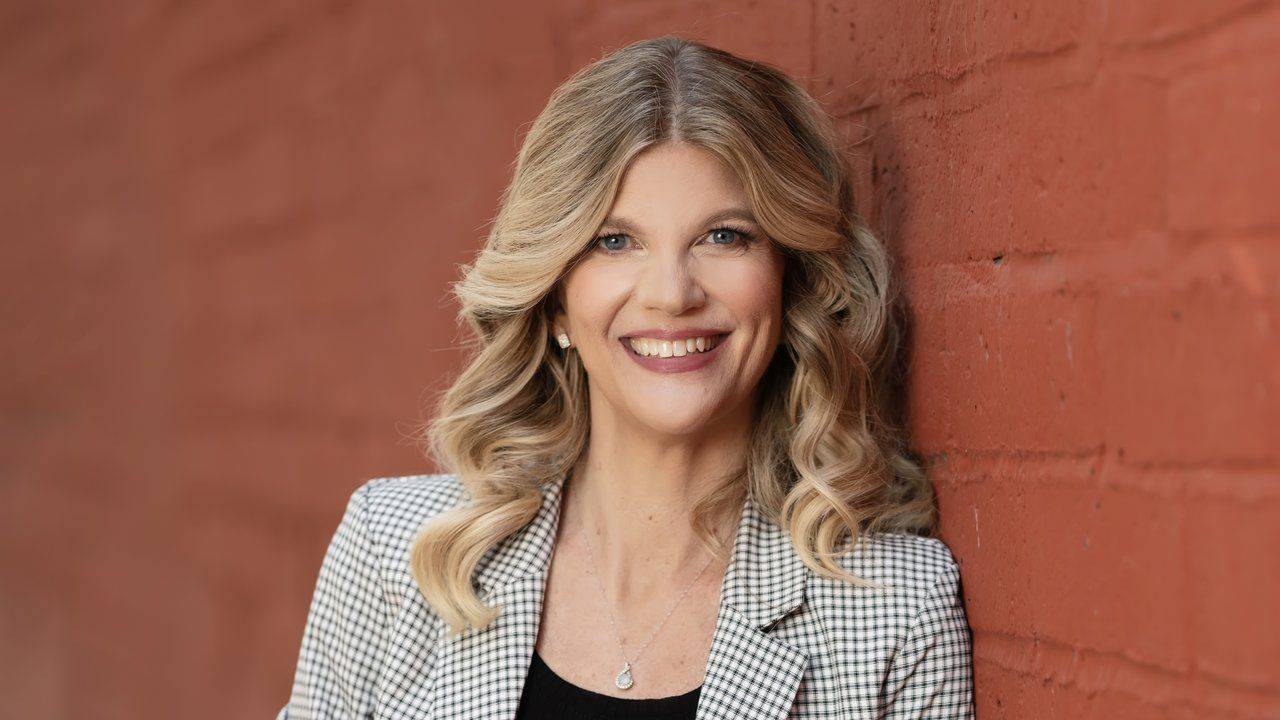 Stacey Salyer smiling in a houndstooth blazer and black top, leaning against a red brick wall