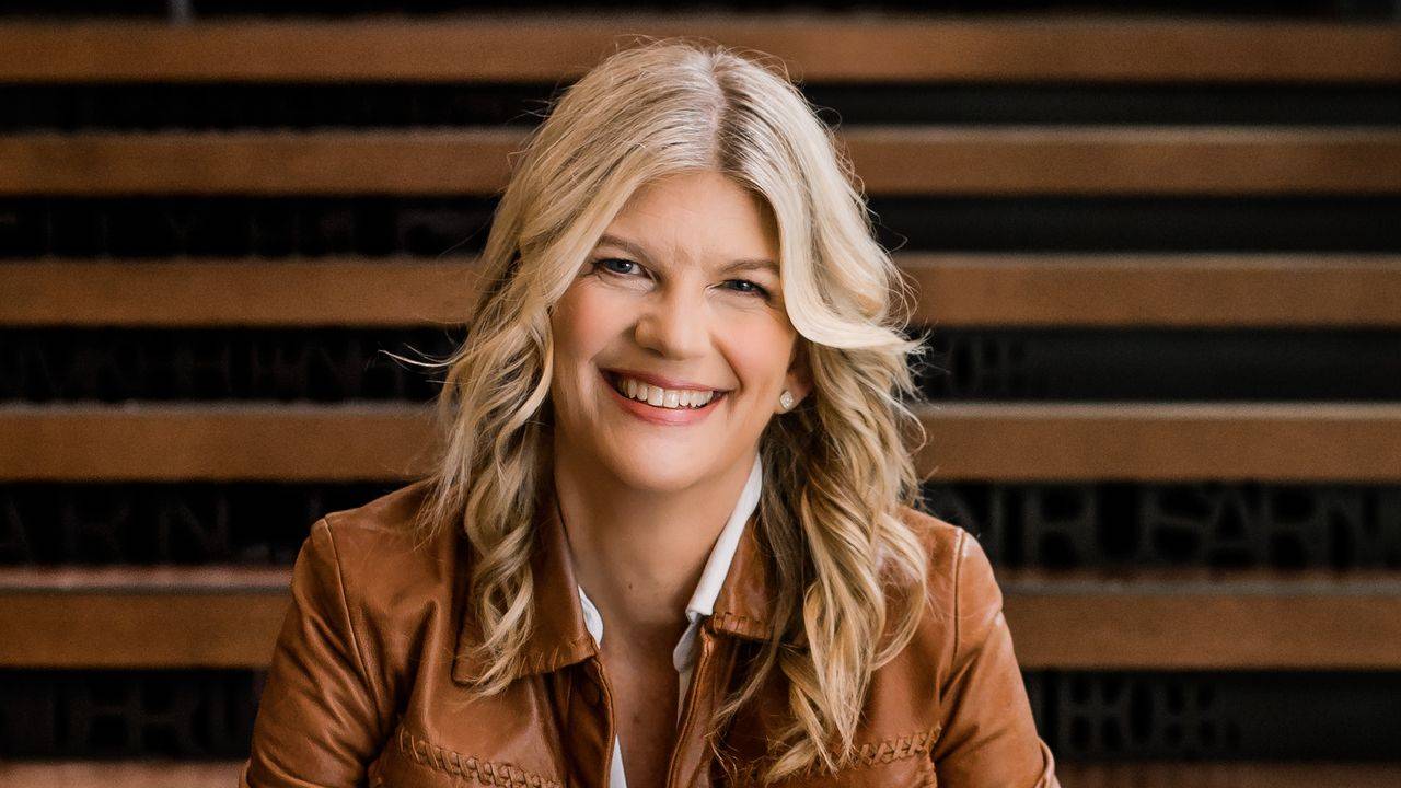 Stacey Salyer smiling in a tan leather jacket, seated on wooden stairs