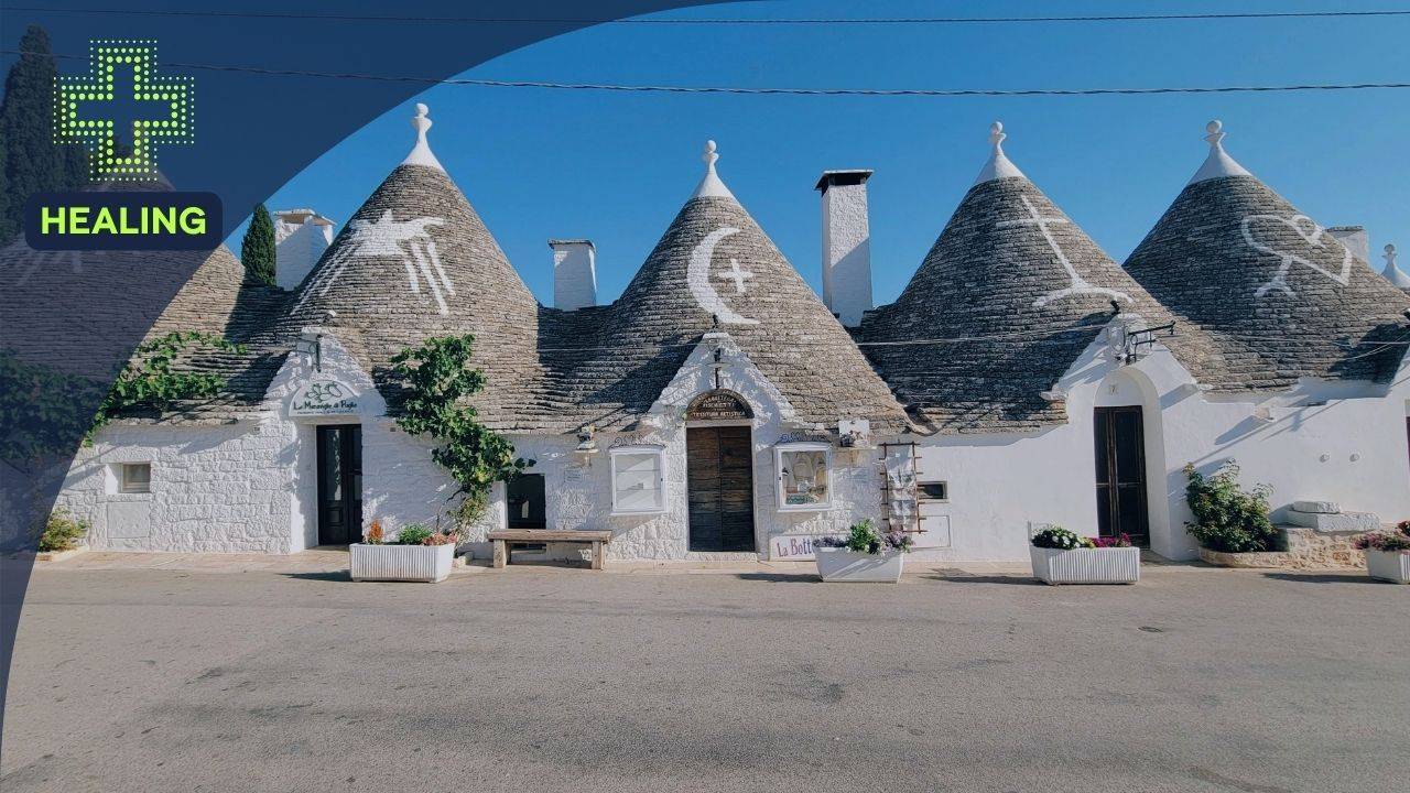 Traditional white Italian trulli buildings with grey conical roofs bearing painted white symbols, situated on a paved street under a clear blue sky, with a 'HEALING' logo in the top left corner.