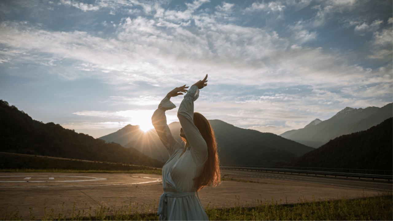 A woman with her hands high with a rising sun among the mountains in the background