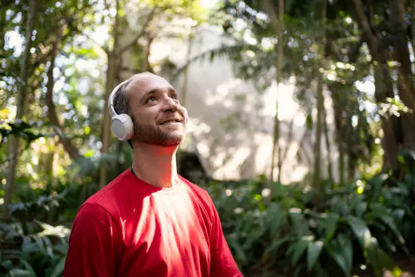 Person in bright red shirt with white headphones standing in a lush green forest, looking upward as sunlight filters through trees.